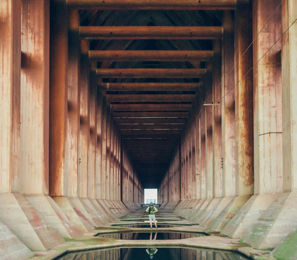 Person with a green backpack standing inside a large, symmetrical concrete tunnel with water pools on the floor.