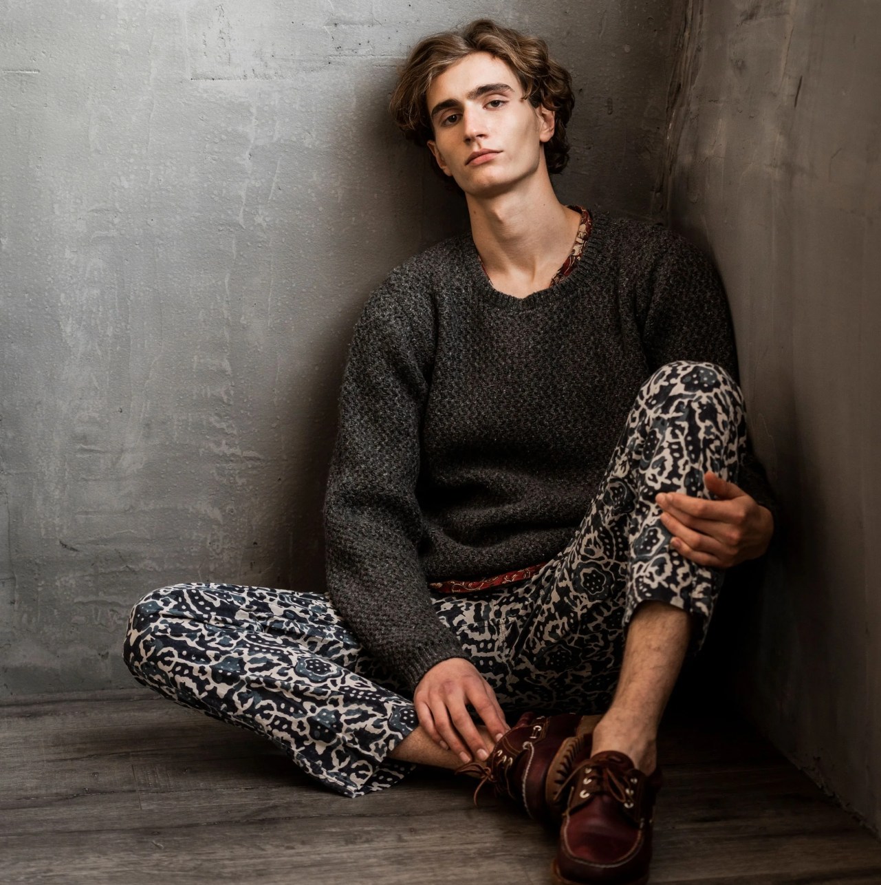 man sitting in a corner while wearing a gray sweater and patterned pants