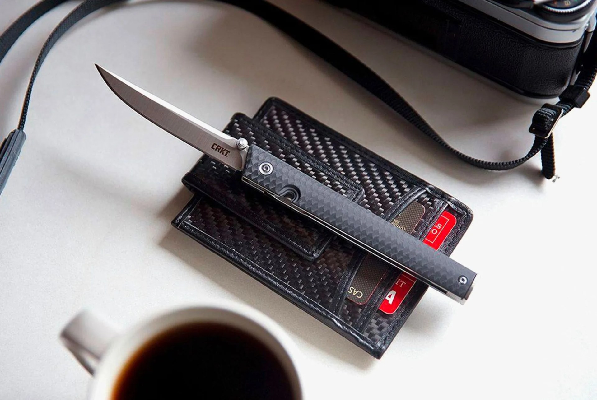 a slim folding pocket knife atop a black wallet on a table next to a cup of coffee