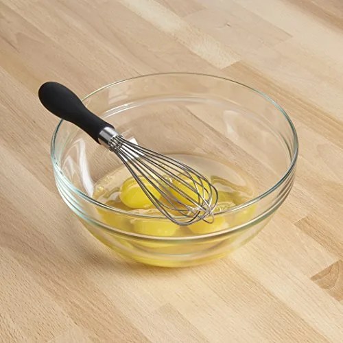 A whisk with a bowl with broken eggs on a wood surface.