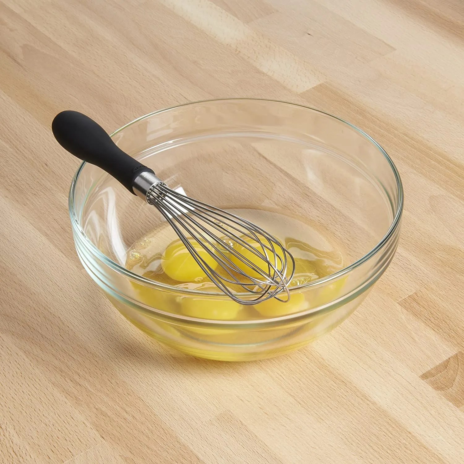 a whisk with a clear bowl with broken egg yolks in it on a wood tabletop
