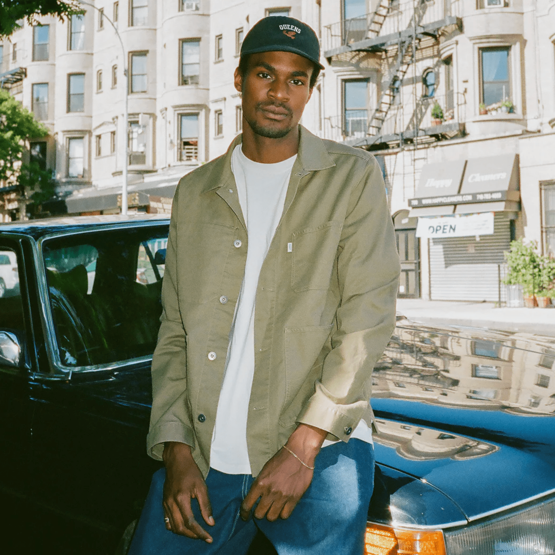 a model sitting on a car wearing a black hat, white t shirt, olive chore coat and blue jeans