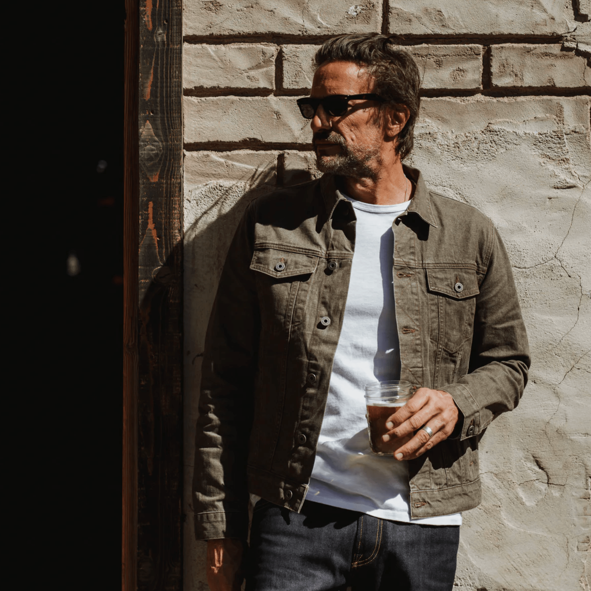a model wearing a brown denim jacket, white t shirt and blue jeans standing against a wall