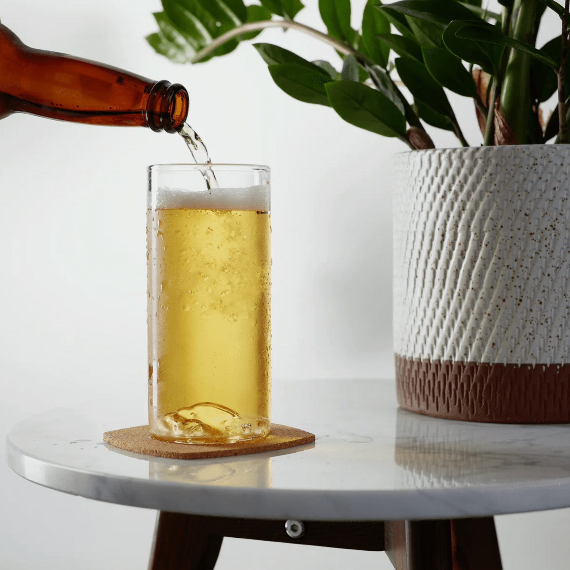 a beer bottle being poured into a beer glass on a table with a plant