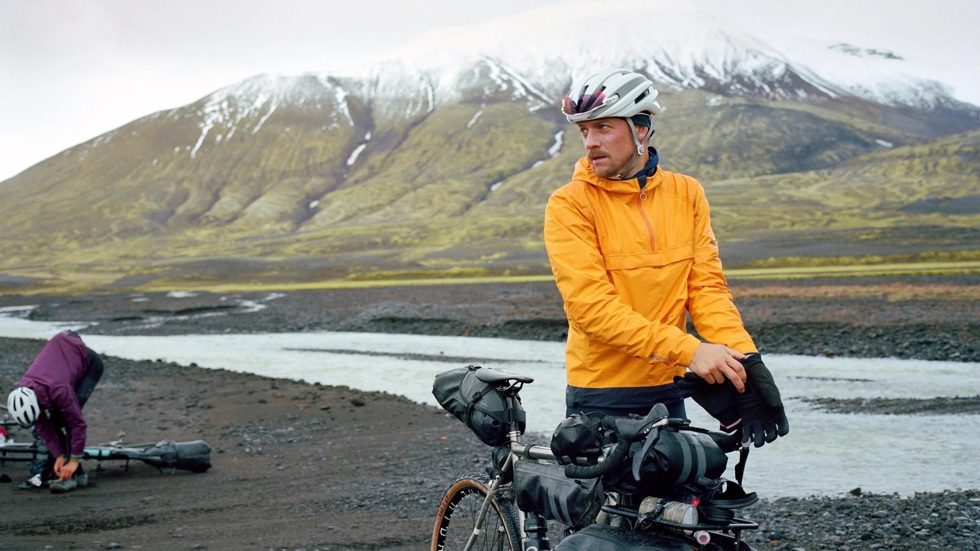 a man wearing an orange pullover standing next to a river and mountain
