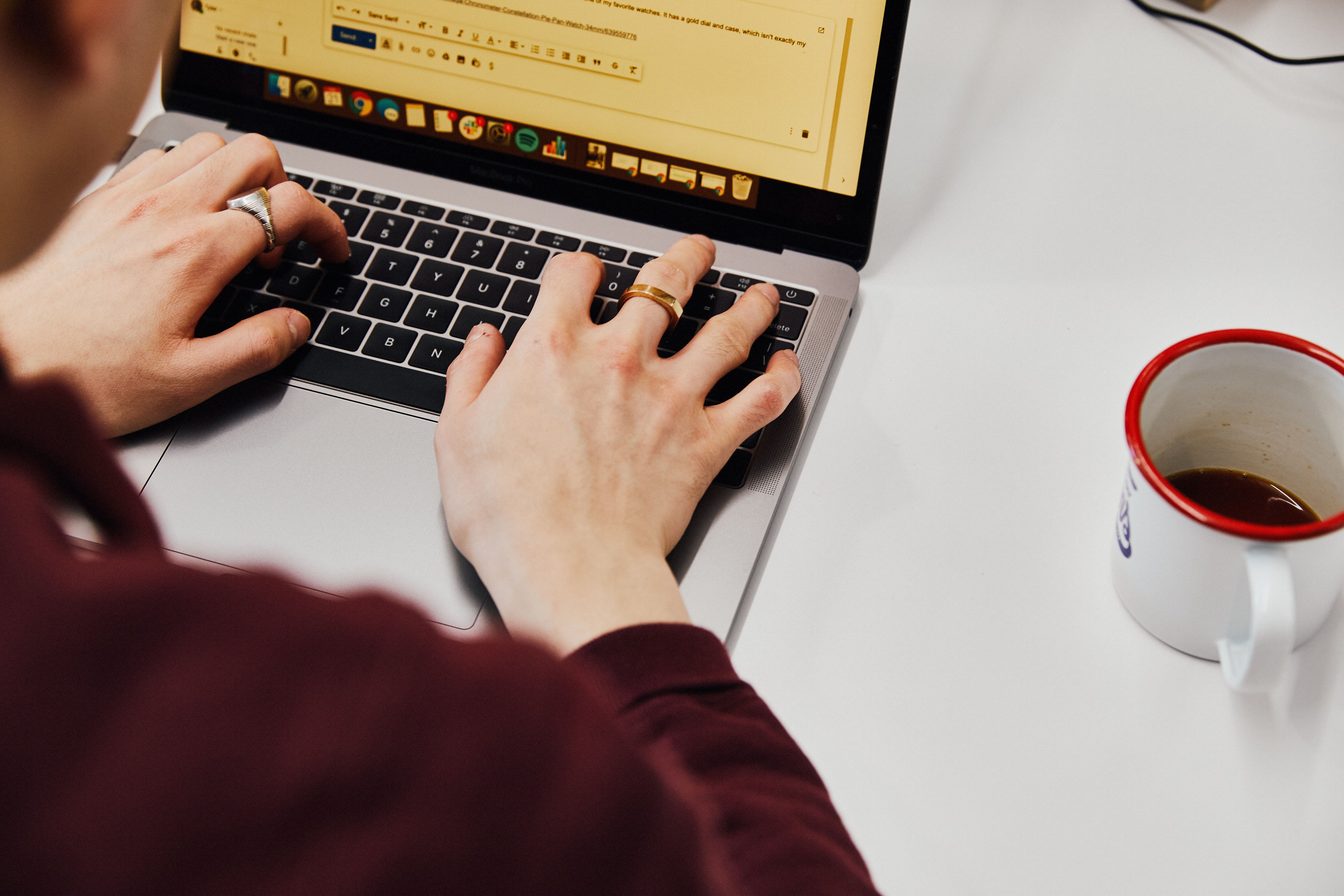 overhead shot of someone typing on a silver macbook on a white table with a cup of coffee to the right of the mabook