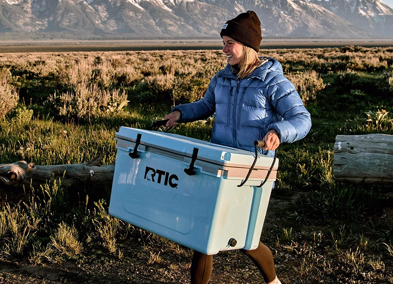 woman carrying rtic ultralight cooler across outdoor landscape with mountains in the background