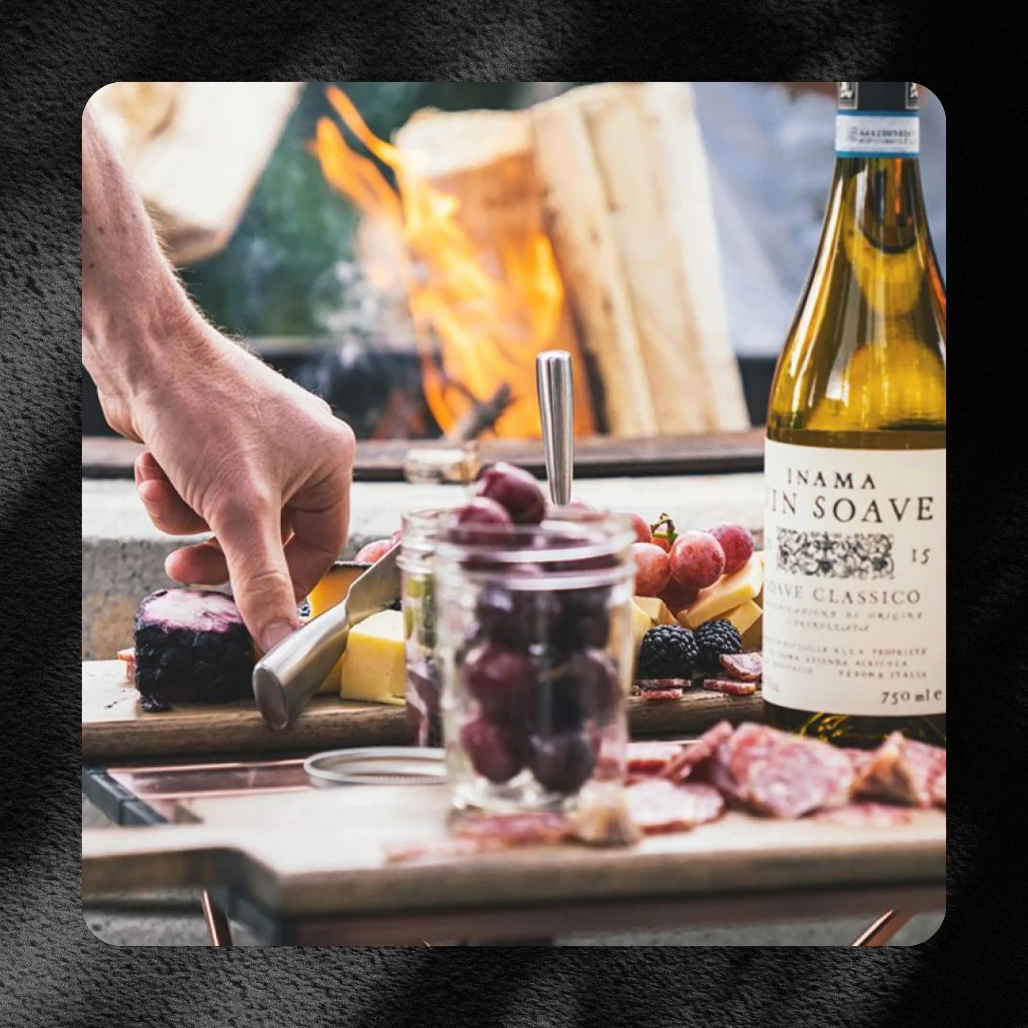 creminelli hosting a party with a hand reaching toward a cured meat on table next to fruit and wine with a small fire in a pit burning in the back