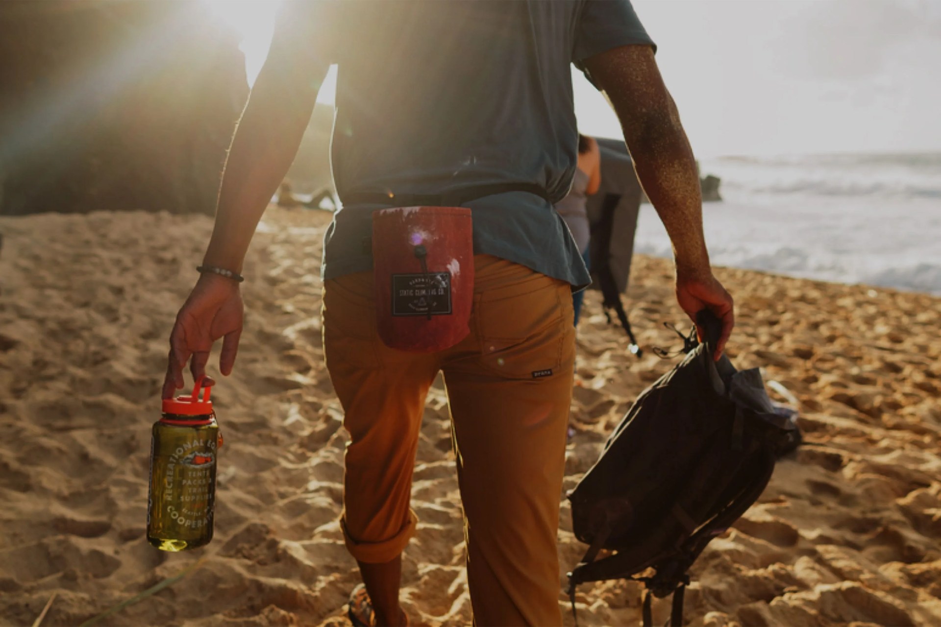 person holding a water bottle and backpack walking on a beach