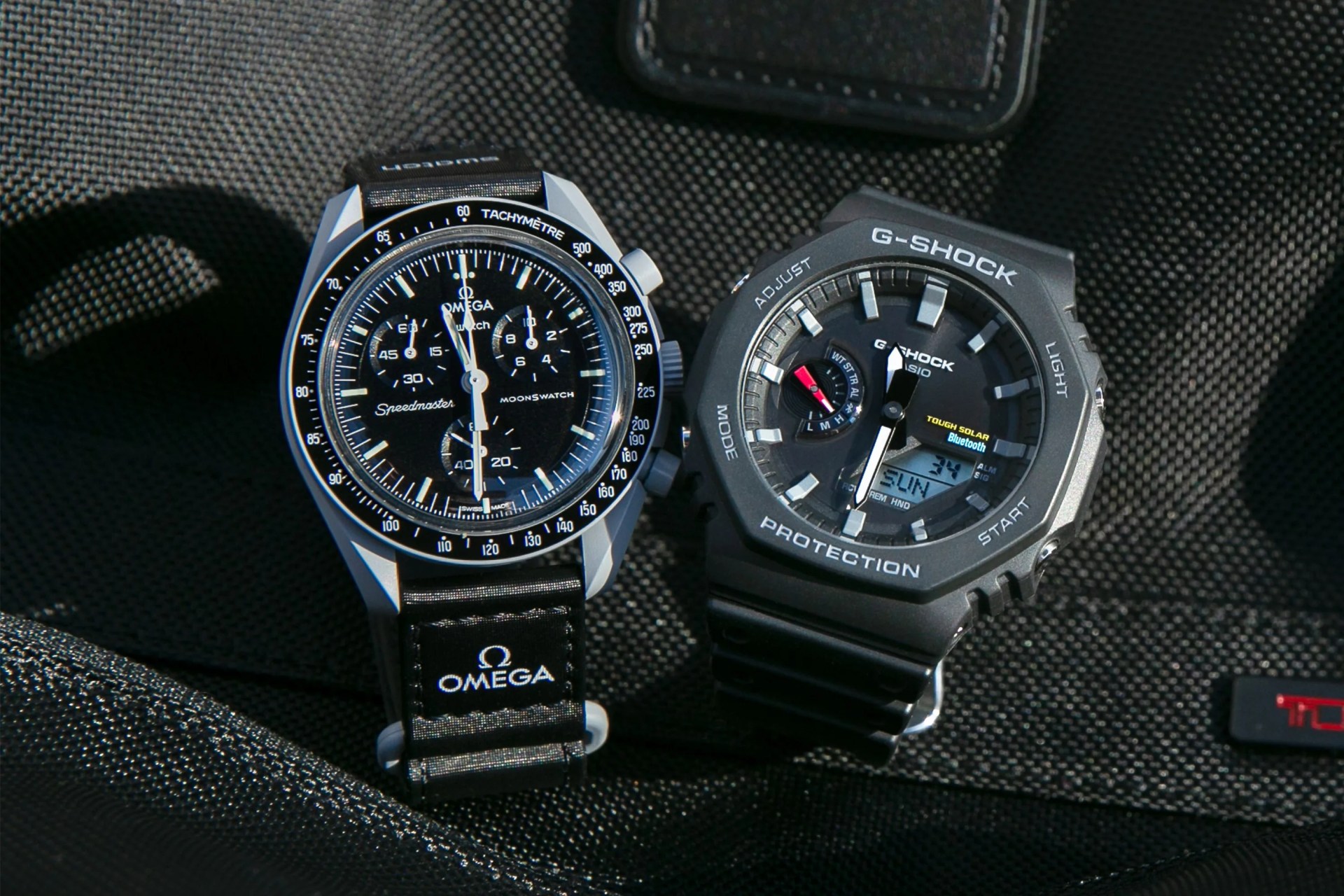 an omega watch and g shock watch next to each other on a black fabric background