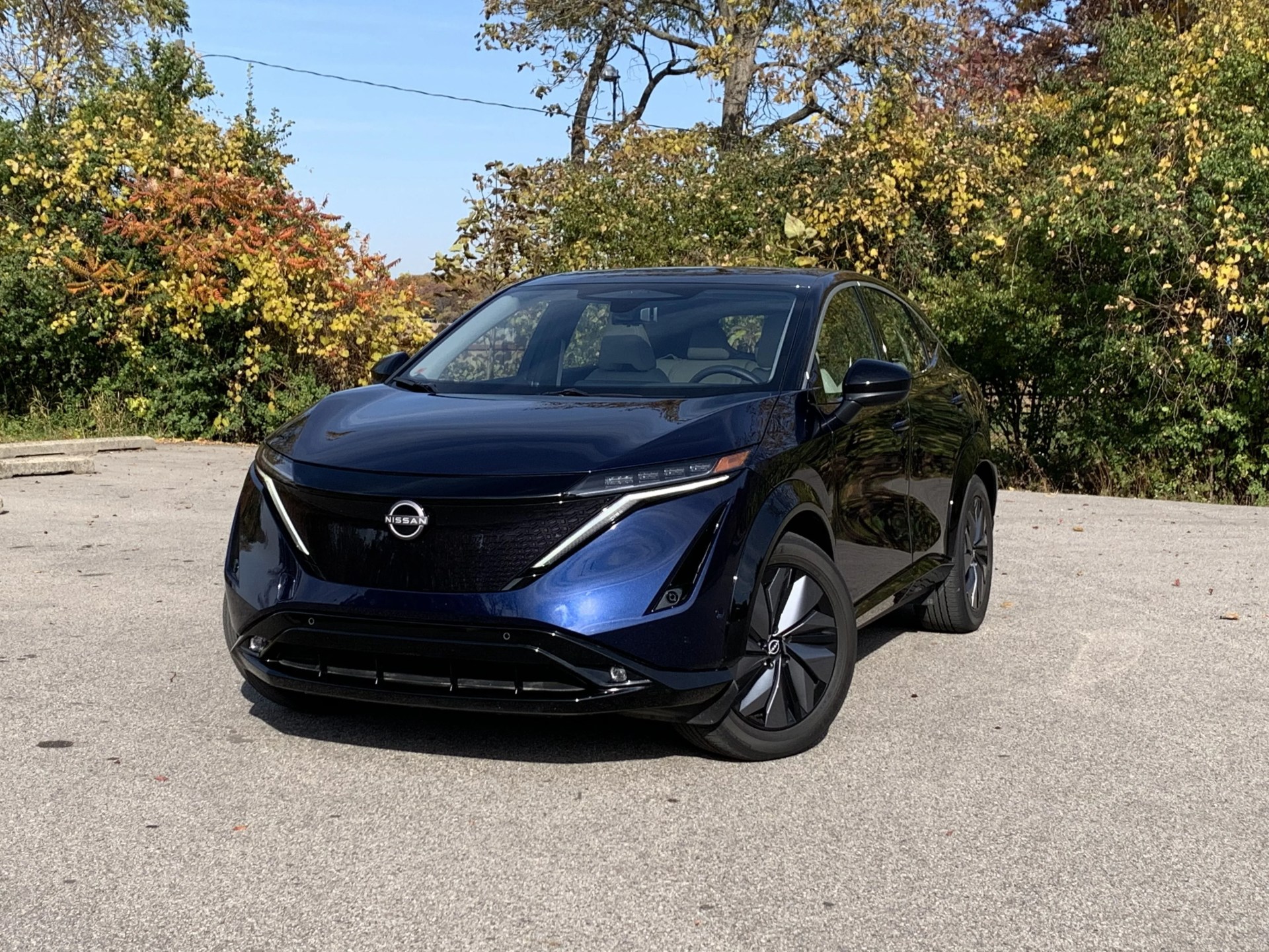 blue 2023 nissan ariya parked on pavement in front of fall foliage