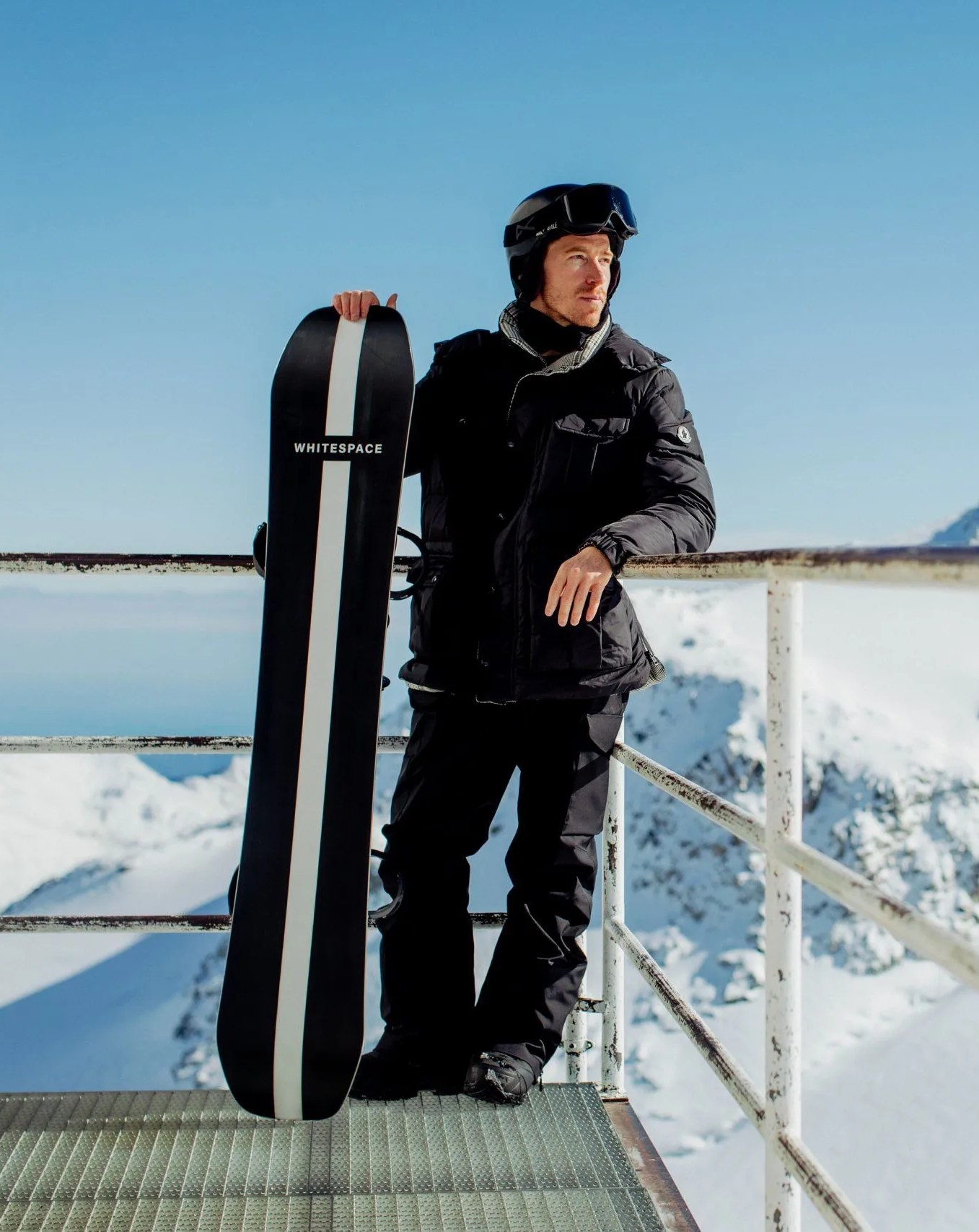 shaun white holding whitespace aspenx freestyle pro snowboard while leaning against railing looking towards the snowy mountains