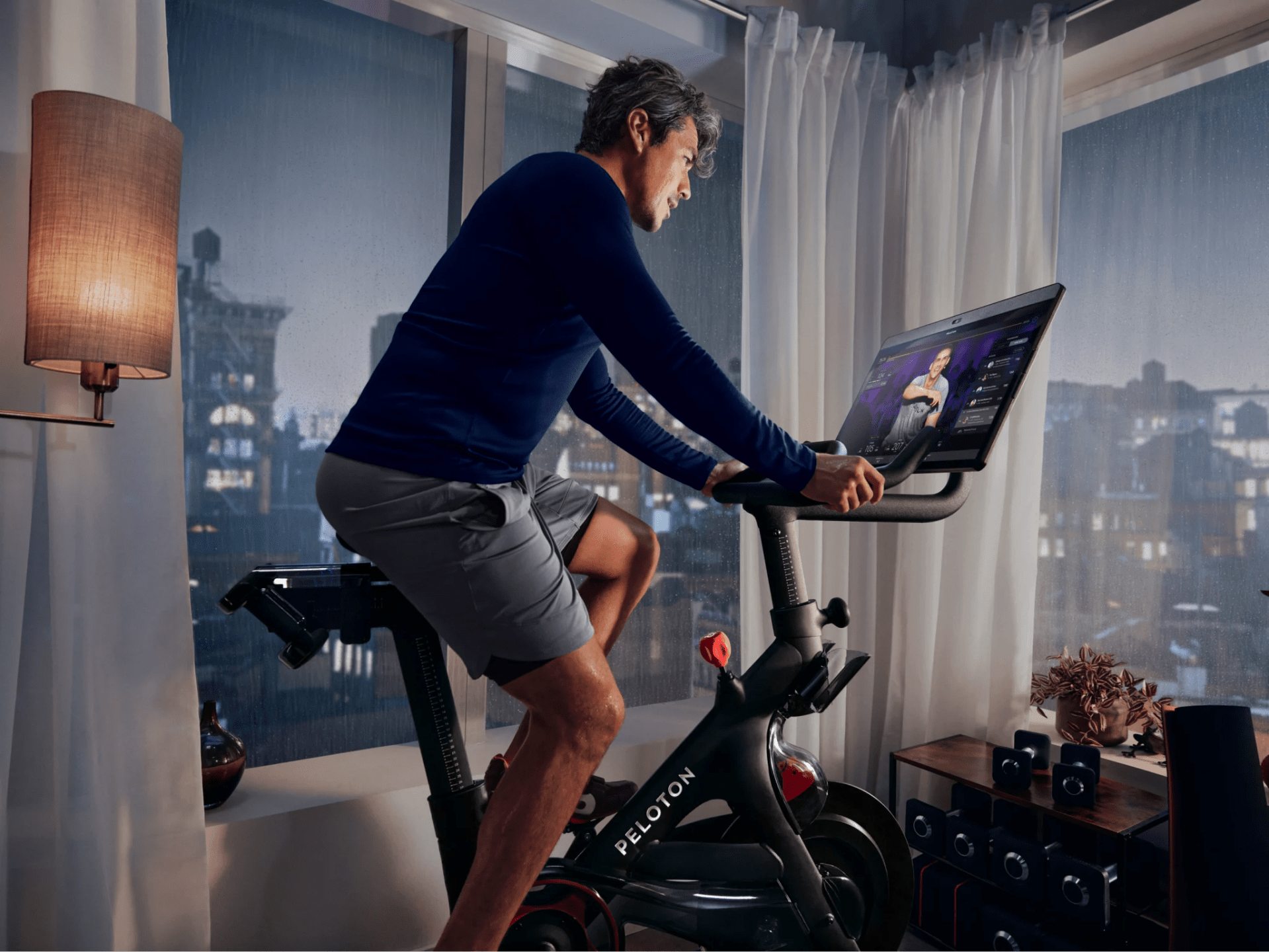 man riding peloton bike in highrise corner apartment in front of window