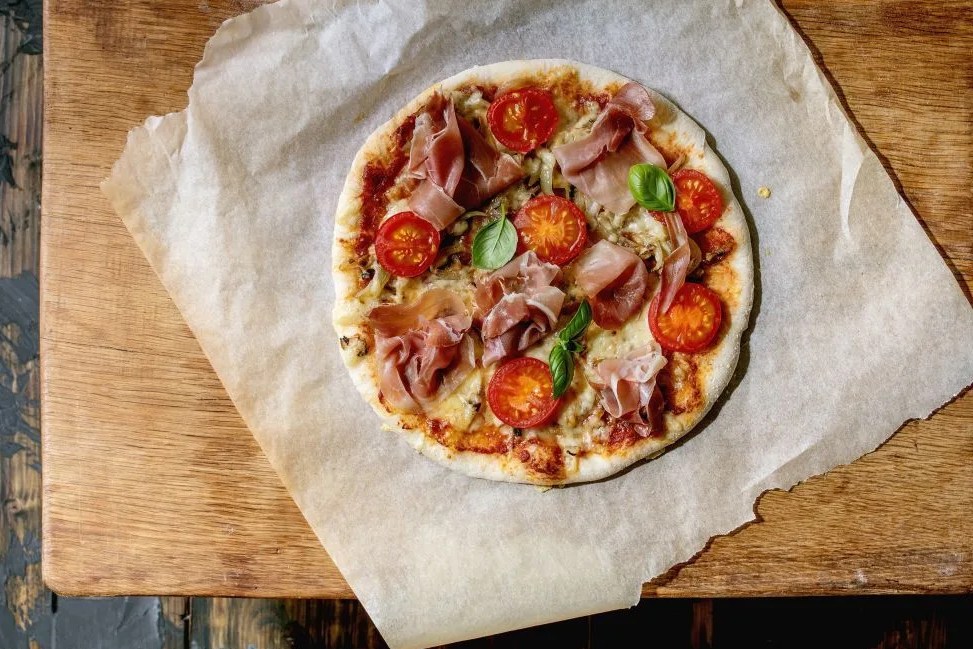 fresh baking homemade pizza napolitana with prosciutto ham, cheese, tomatoes, basil on baking paper over wooden table background home baking or delivered fast food flat lay, space