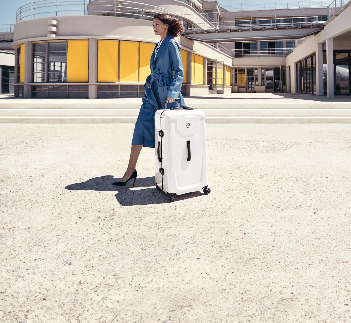 a woman in a blue coat pushing a white peugeot voyages suitcase