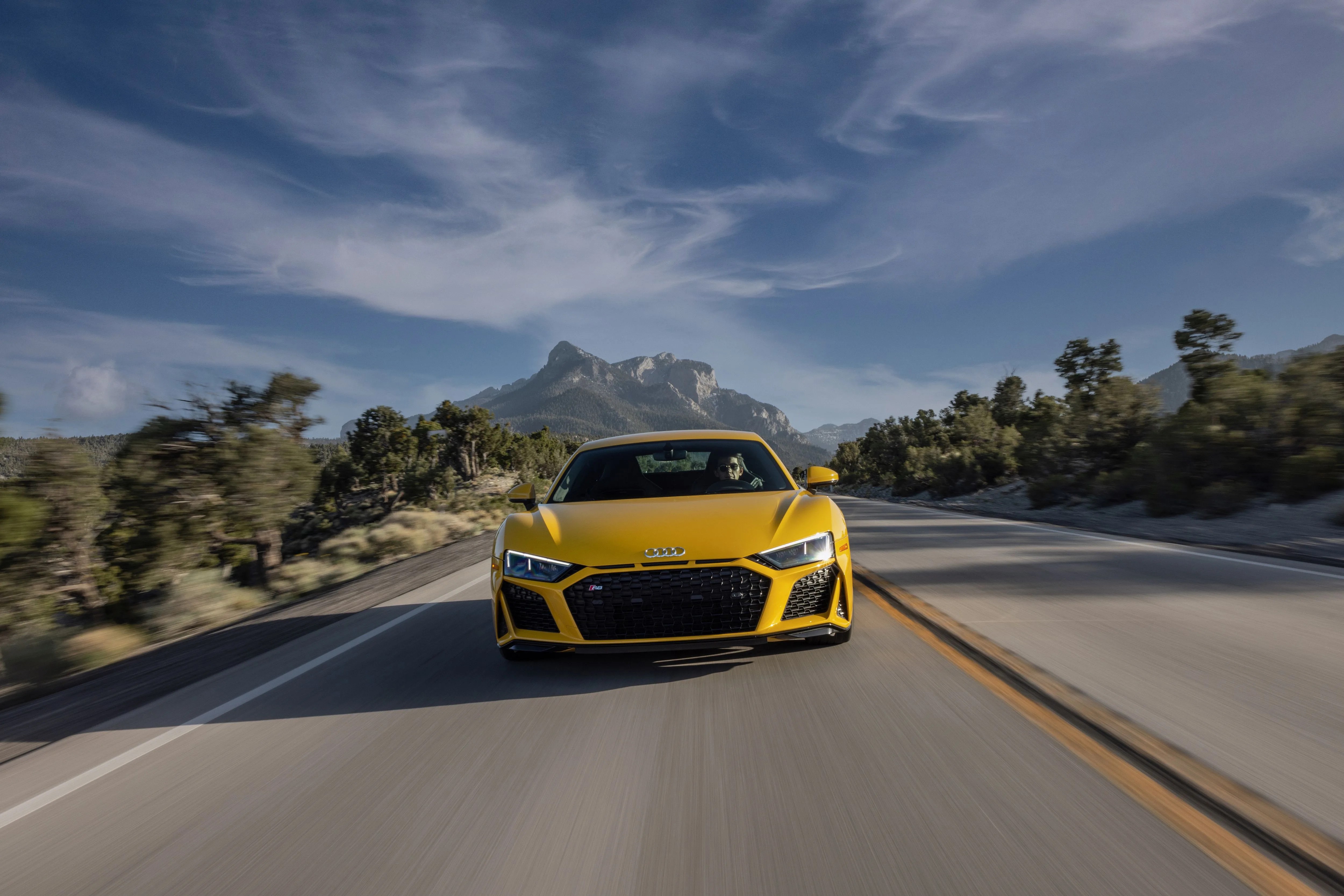 a yellow sports car driving on a road with trees and mountains in the background