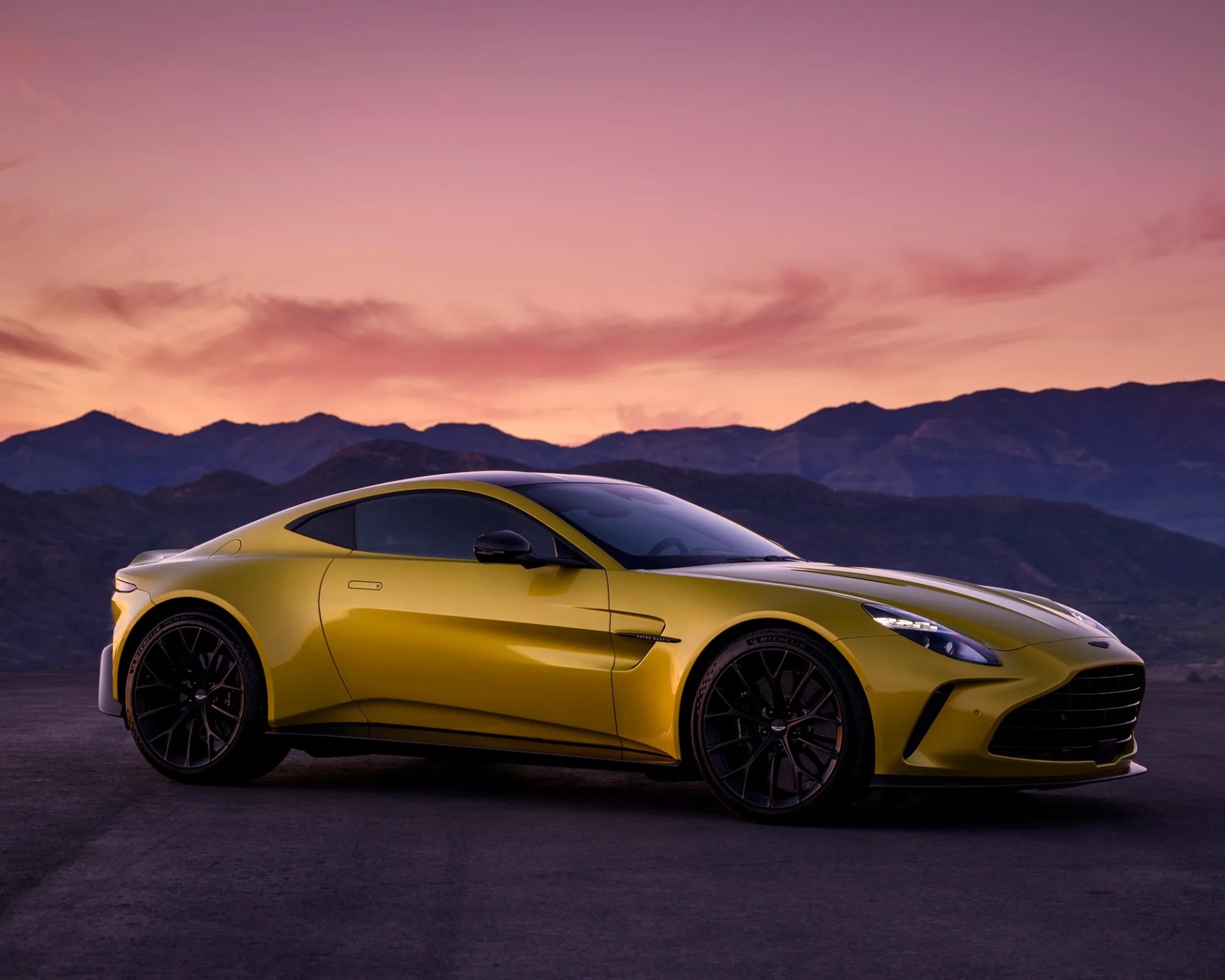 a gold aston martin vantage parked on a hill in front of mountains at sunset