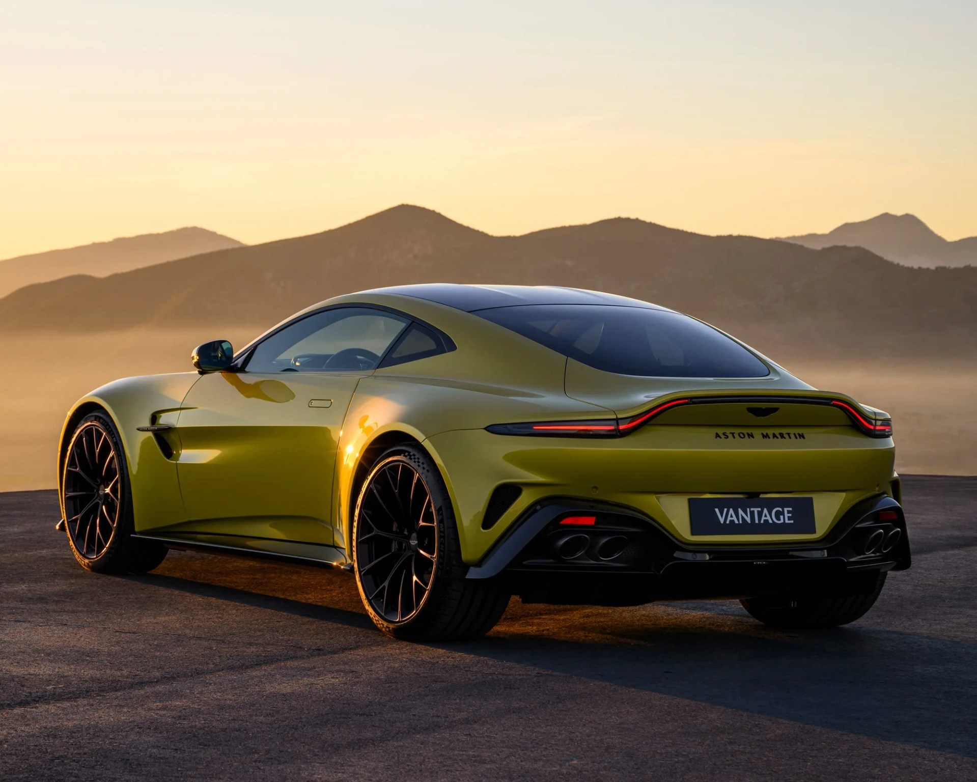 a gold aston martin vantage parked on a hill in front of mountains at sunset