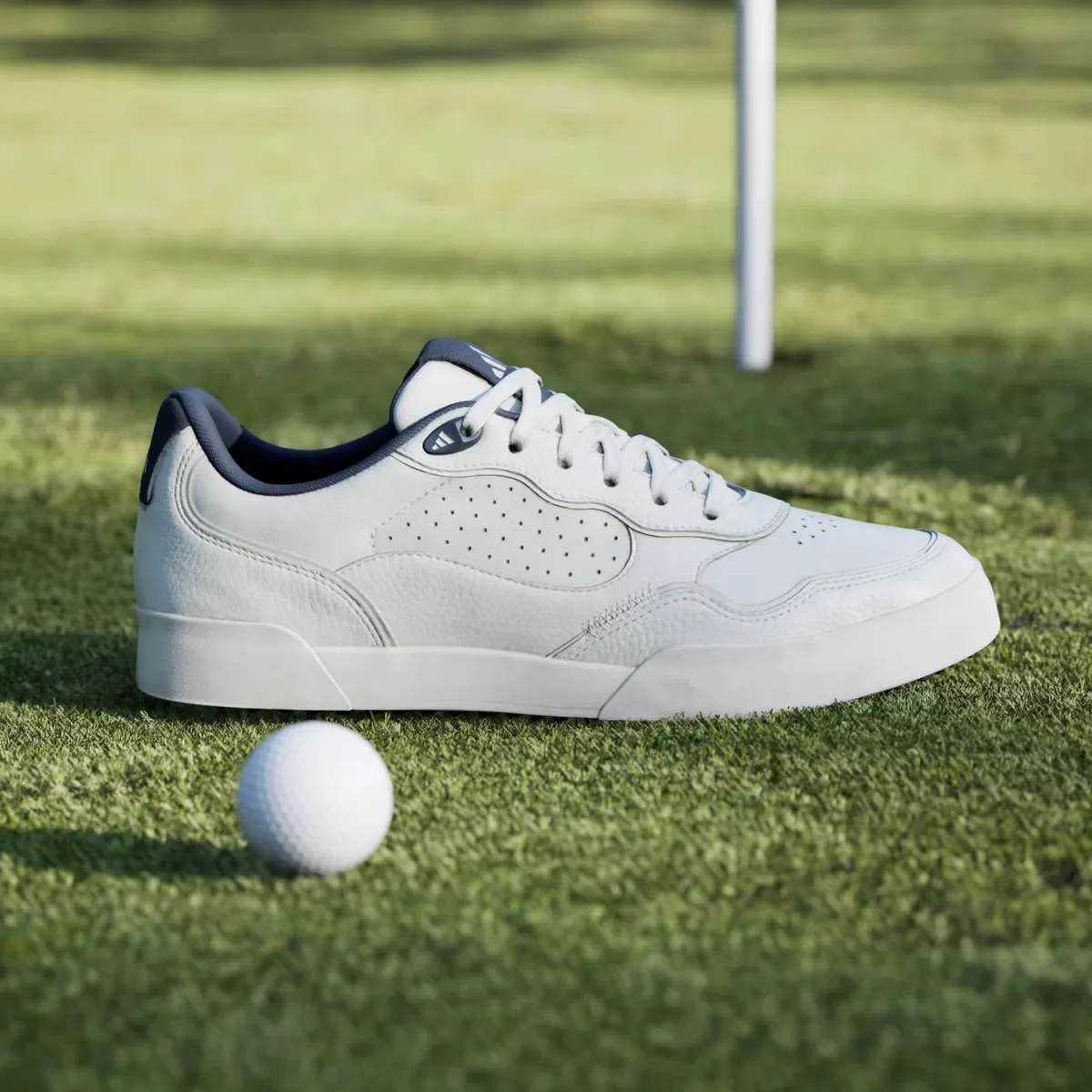 White leather sneaker with navy lining and laces on grass next to a golf ball.