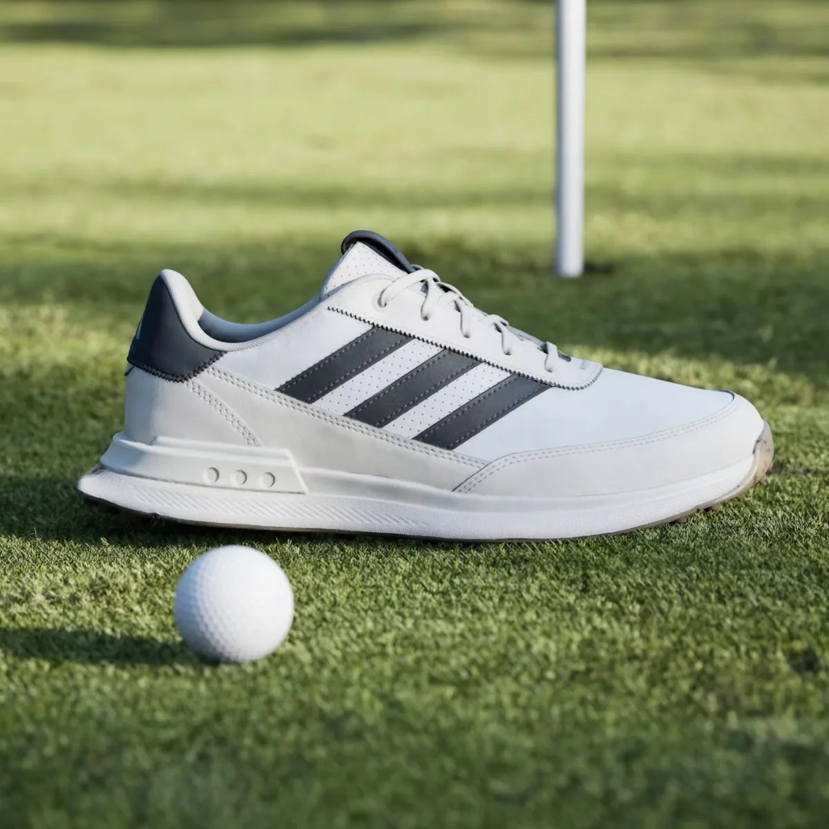 White and black Adidas golf shoe on grass next to a golf ball and flagstick.