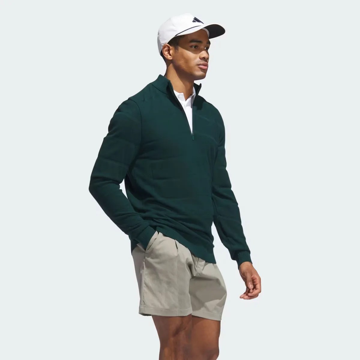 Man wearing dark green quarter-zip pullover, beige shorts, white polo, and white Adidas cap.