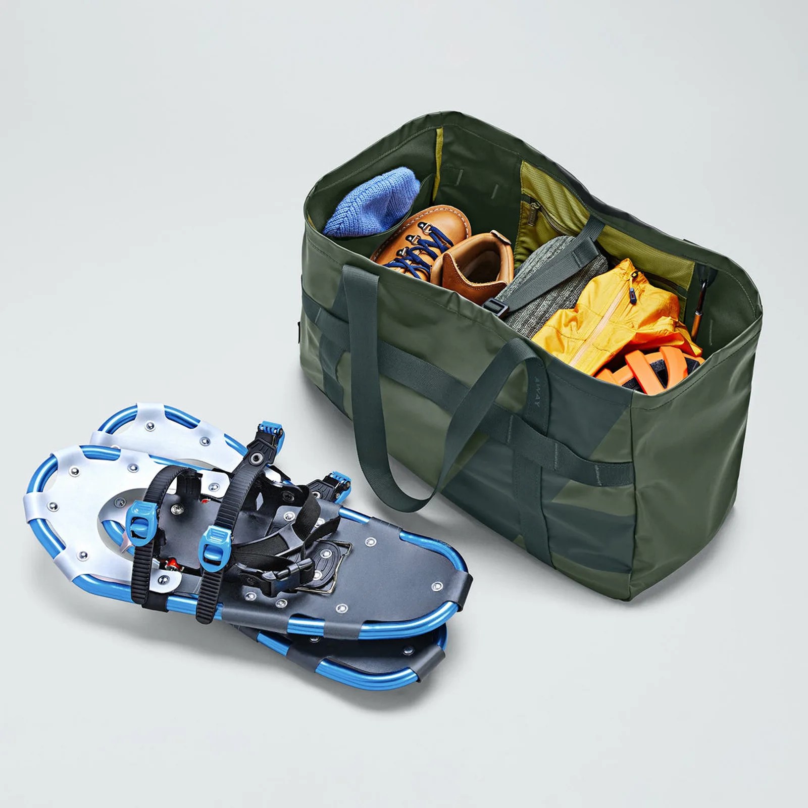 A dark green duffel bag filled with outdoor gear including brown hiking boots, a blue knit item, a gray sweater, a yellow jacket, and orange straps. Next to the bag is a pair of blue and black snowshoes with adjustable straps.