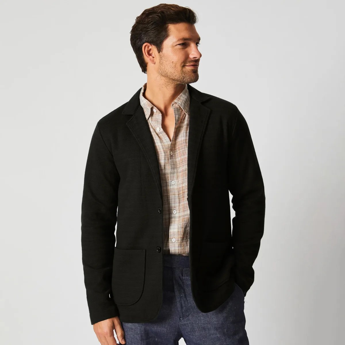Man wearing a black blazer over a beige plaid button-up shirt and blue pants against a plain background.