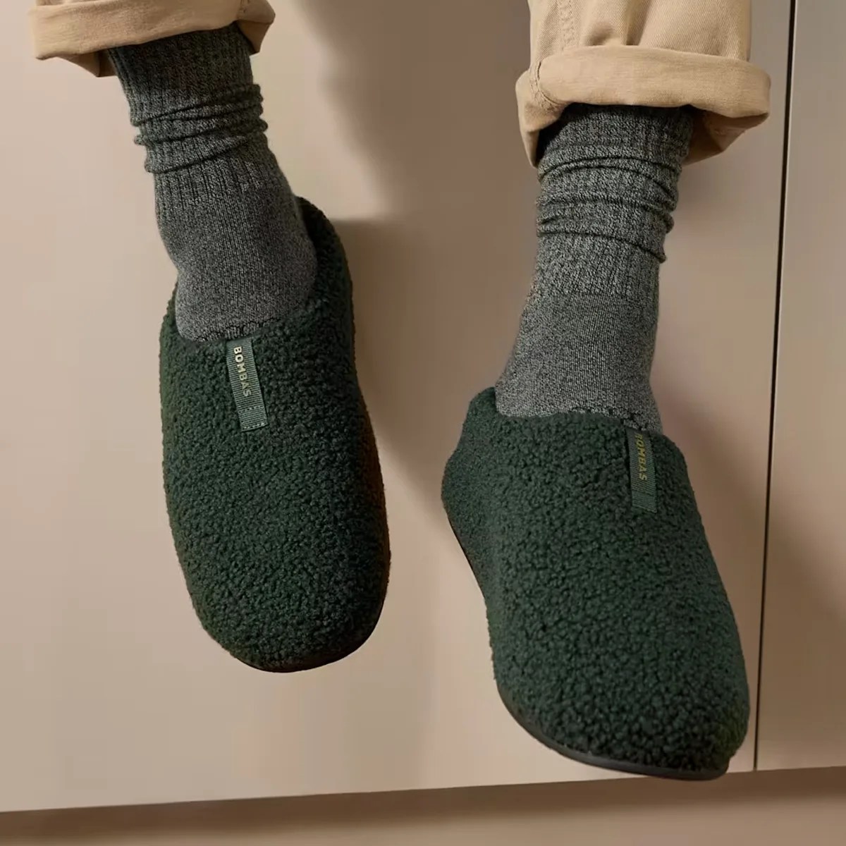 Dark green textured slip-on slippers worn with gray socks and beige pants.