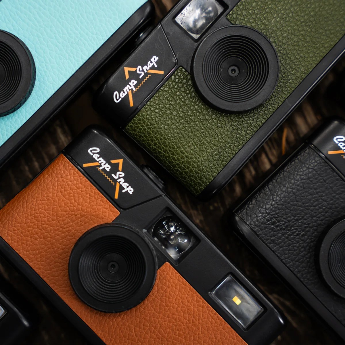 Four Camp Snap cameras with textured leather-like covers in turquoise, olive green, orange, and black.