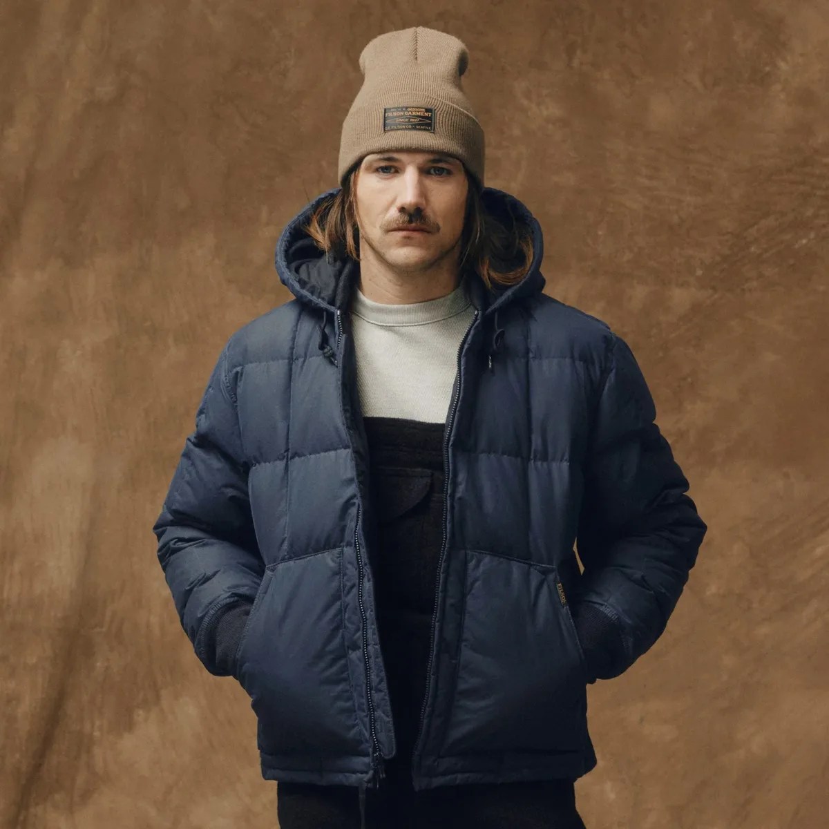 Man wearing a navy blue puffer jacket with a hood and a beige knit beanie with a patch on the front.