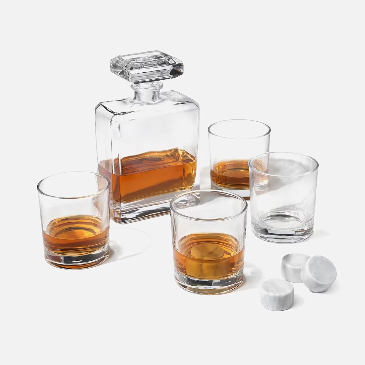 Square glass whiskey decanter with stopper, three whiskey glasses with amber liquid, one empty glass, and four marble whiskey stones.