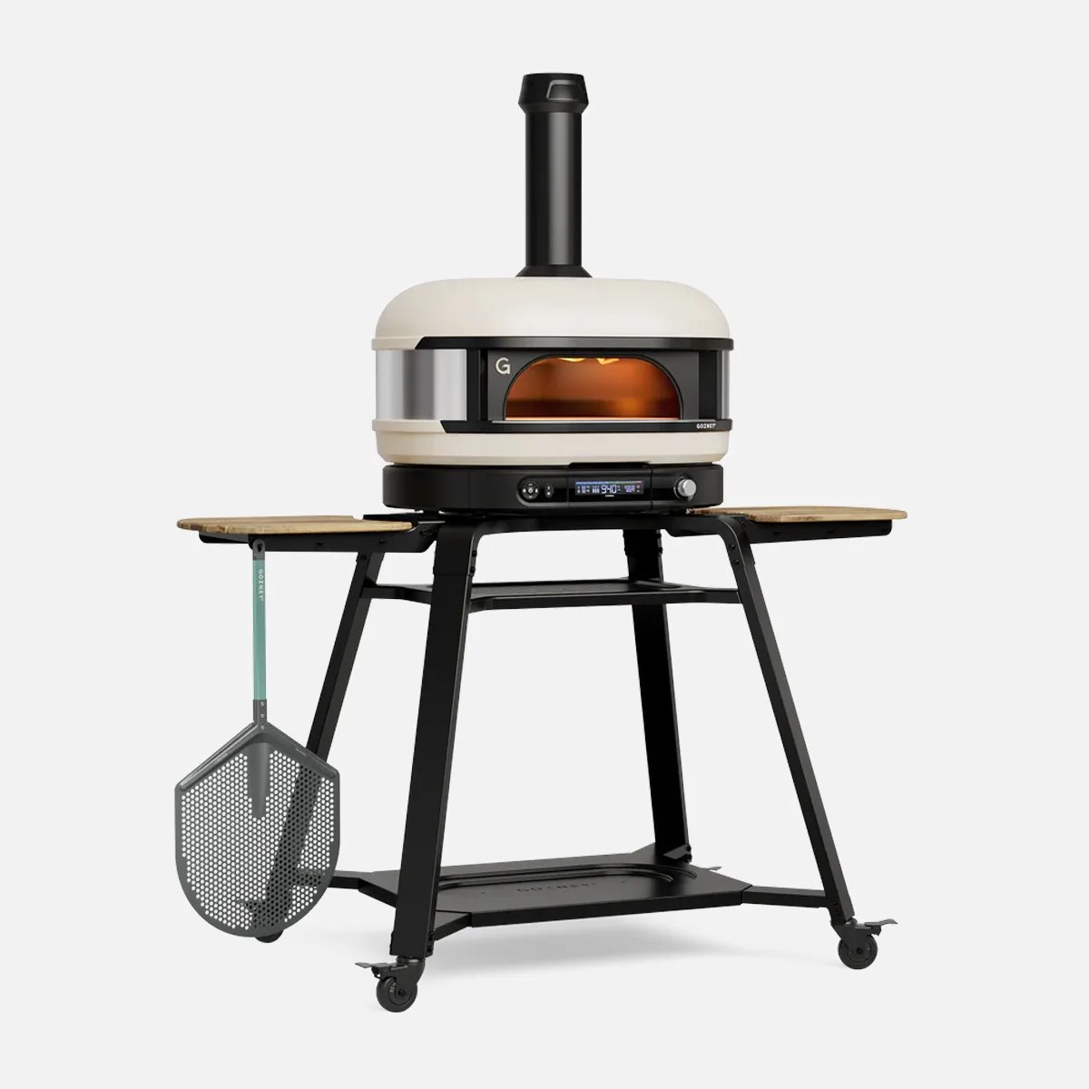 Cream and black outdoor pizza oven on a black metal stand with wooden side shelves and a pizza peel hanging on the side.