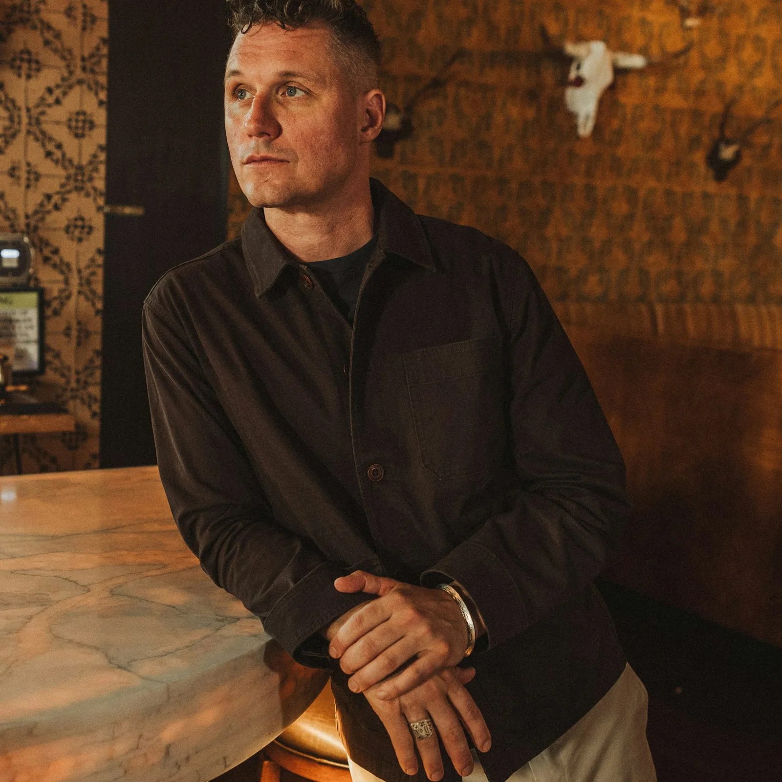 Man wearing a dark button-up shirt leaning on a marble countertop in a warmly lit room.