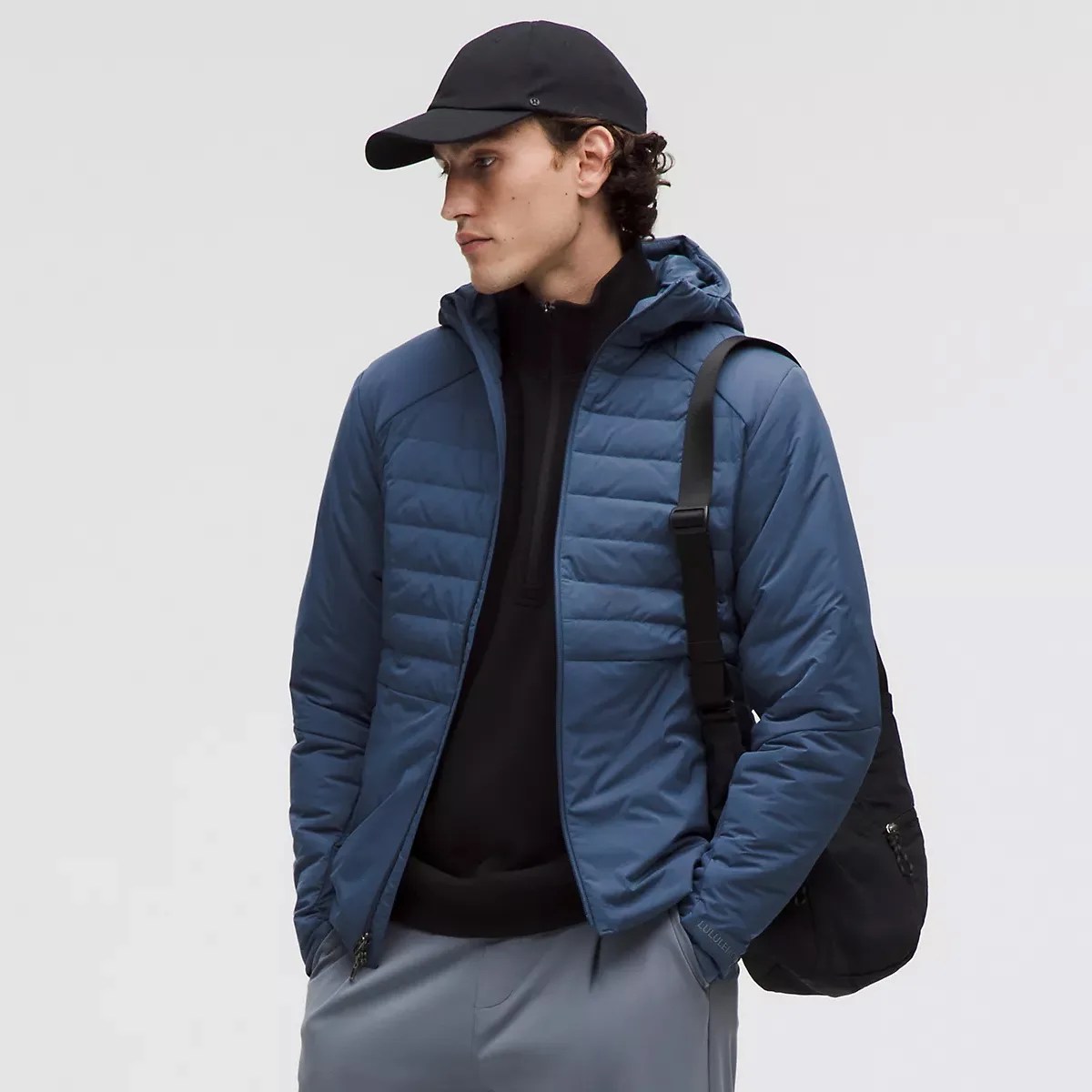 Man wearing a blue quilted hooded jacket over a black zip-up top, gray pants, black cap, and carrying a black shoulder bag.