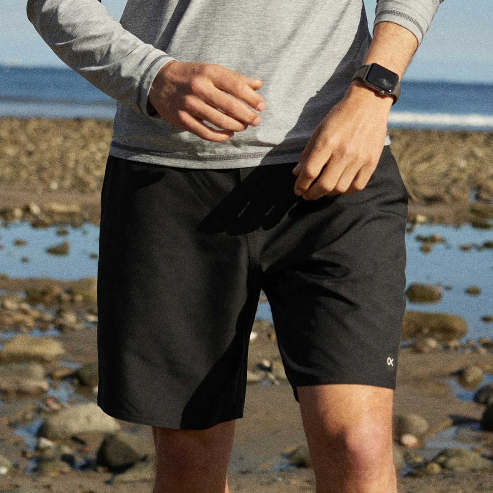 A pair of Outerknown Apex Hybrid shorts in black being worn by a man walking on a rocky beach.