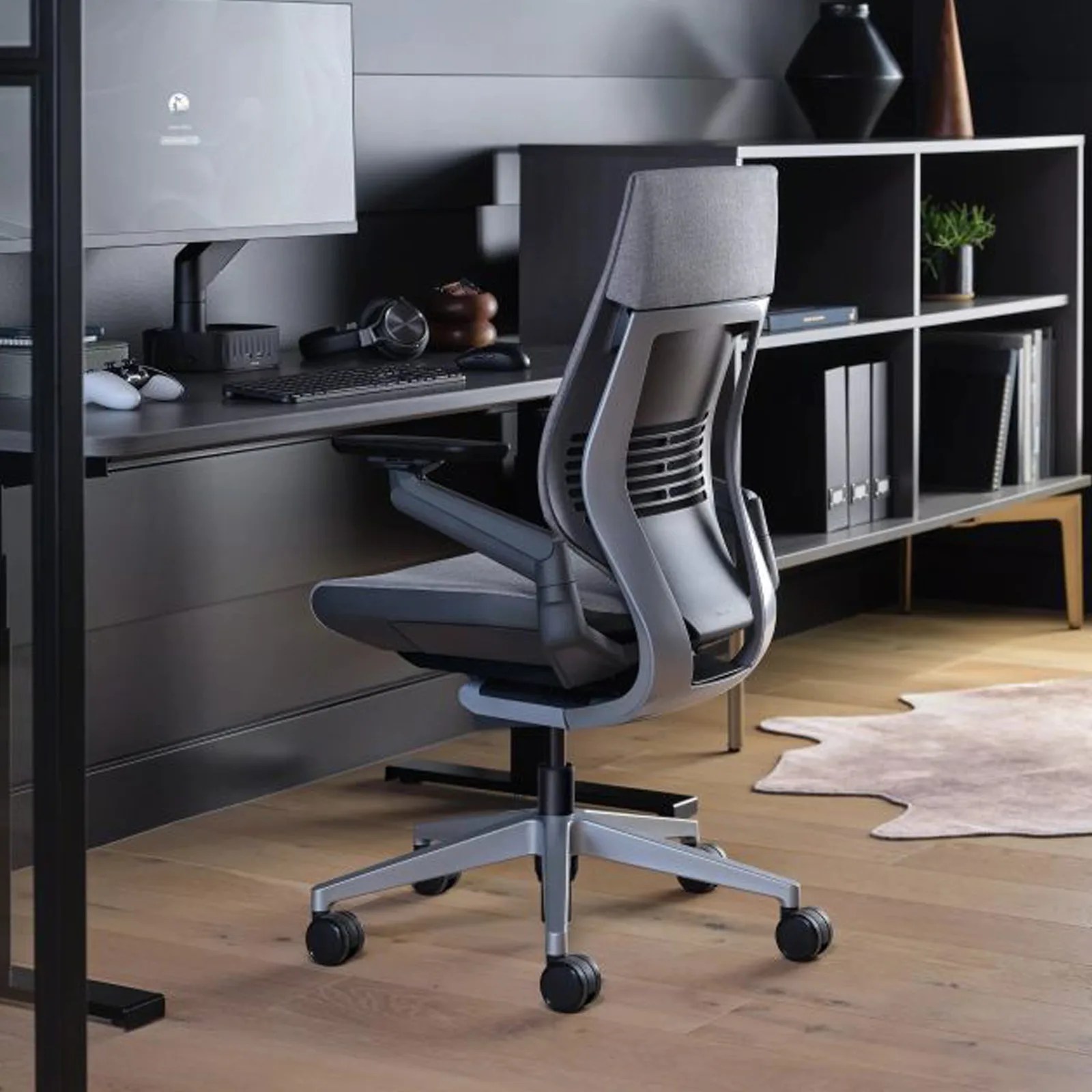Gray ergonomic office chair with headrest and black mesh backrest on wooden floor near black desk and shelving unit.