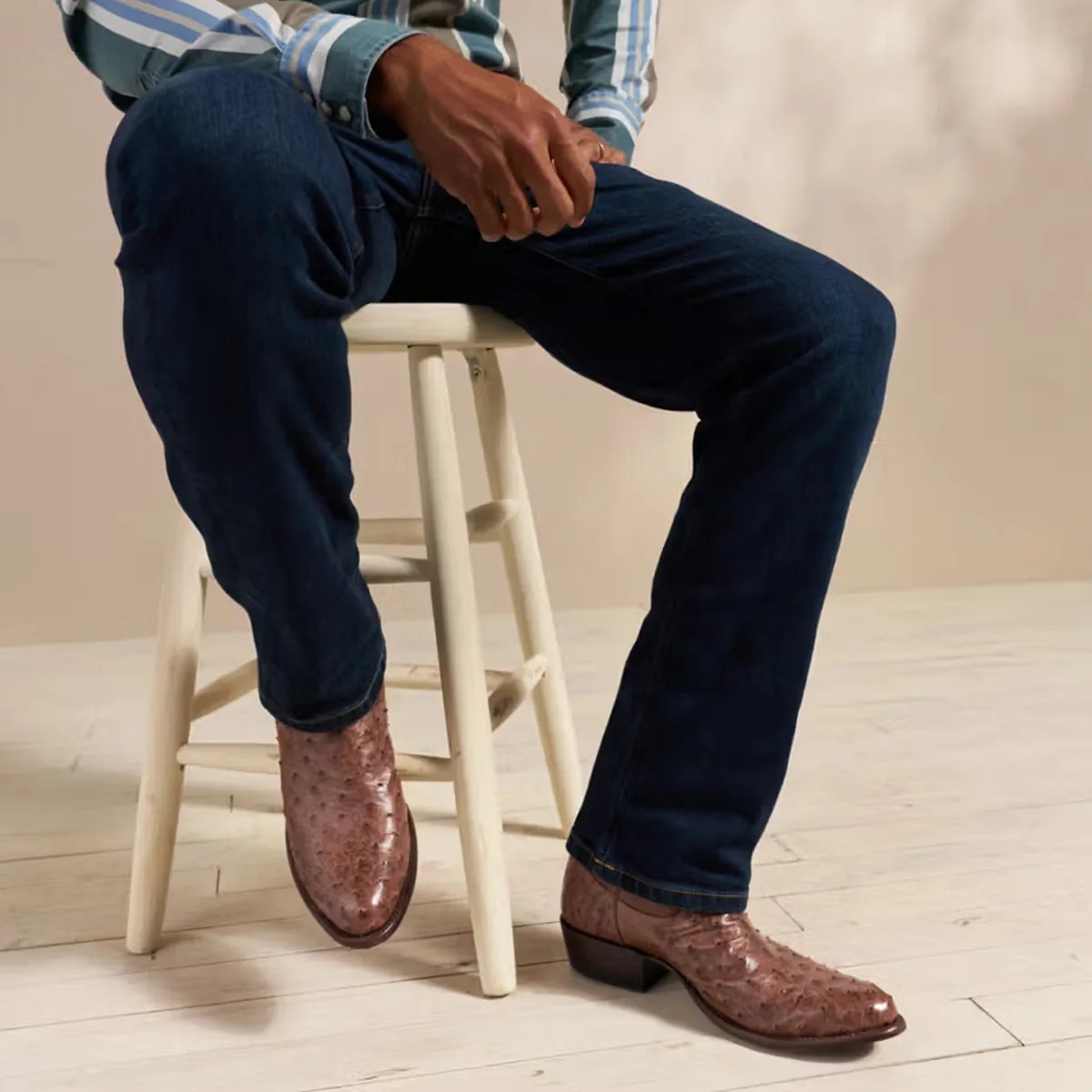 Brown ostrich leather dress shoes worn with dark blue jeans while sitting on a white wooden stool.
