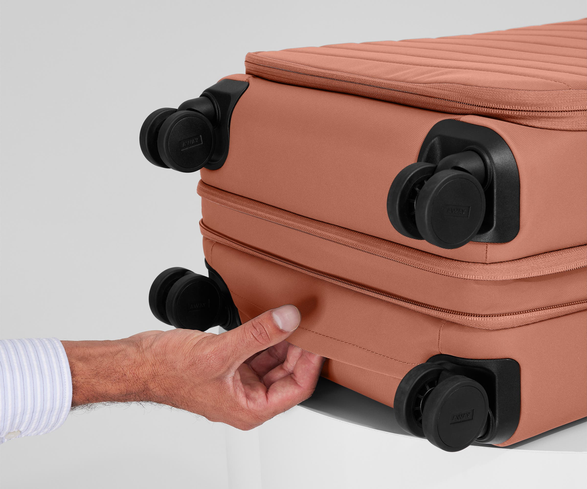 A hand pulling the bottom handle of a salmon-colored version of Away’s Softside carry-on luggage that’s sitting on its back against a grey backdrop.