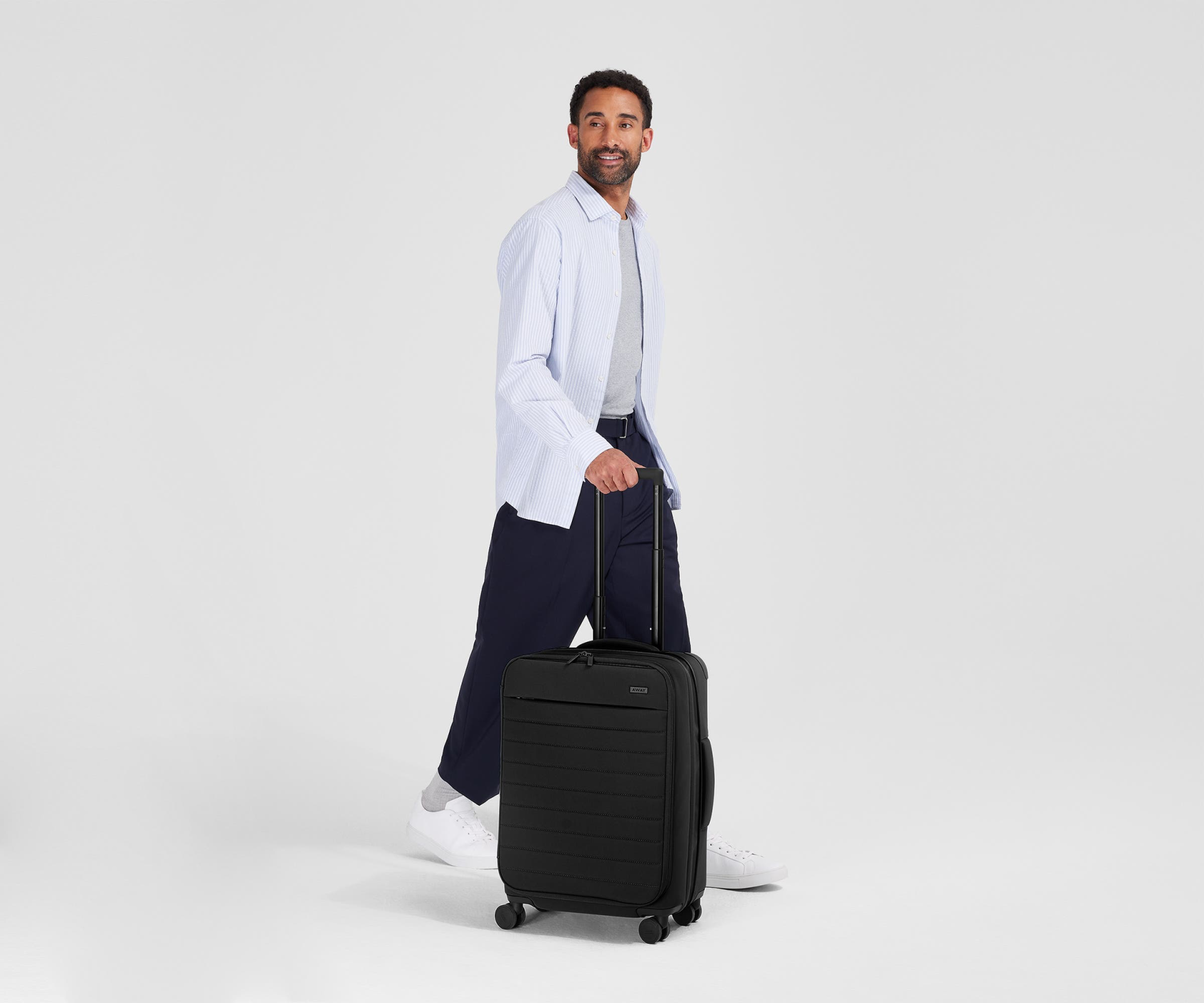 Away Softside carry-on in black being rolled by a man holding the handle and smiling in a blank grey space.  