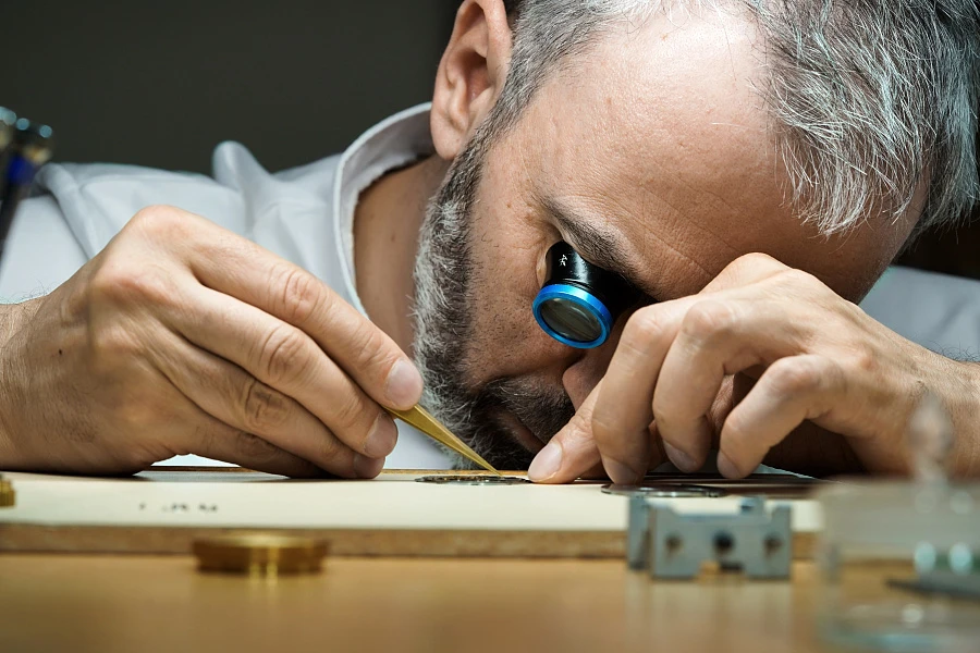 Konstantin Chaykin working on a watch movement using a small watch tool while sitting at a table with a magnifying loupe in one eye.
