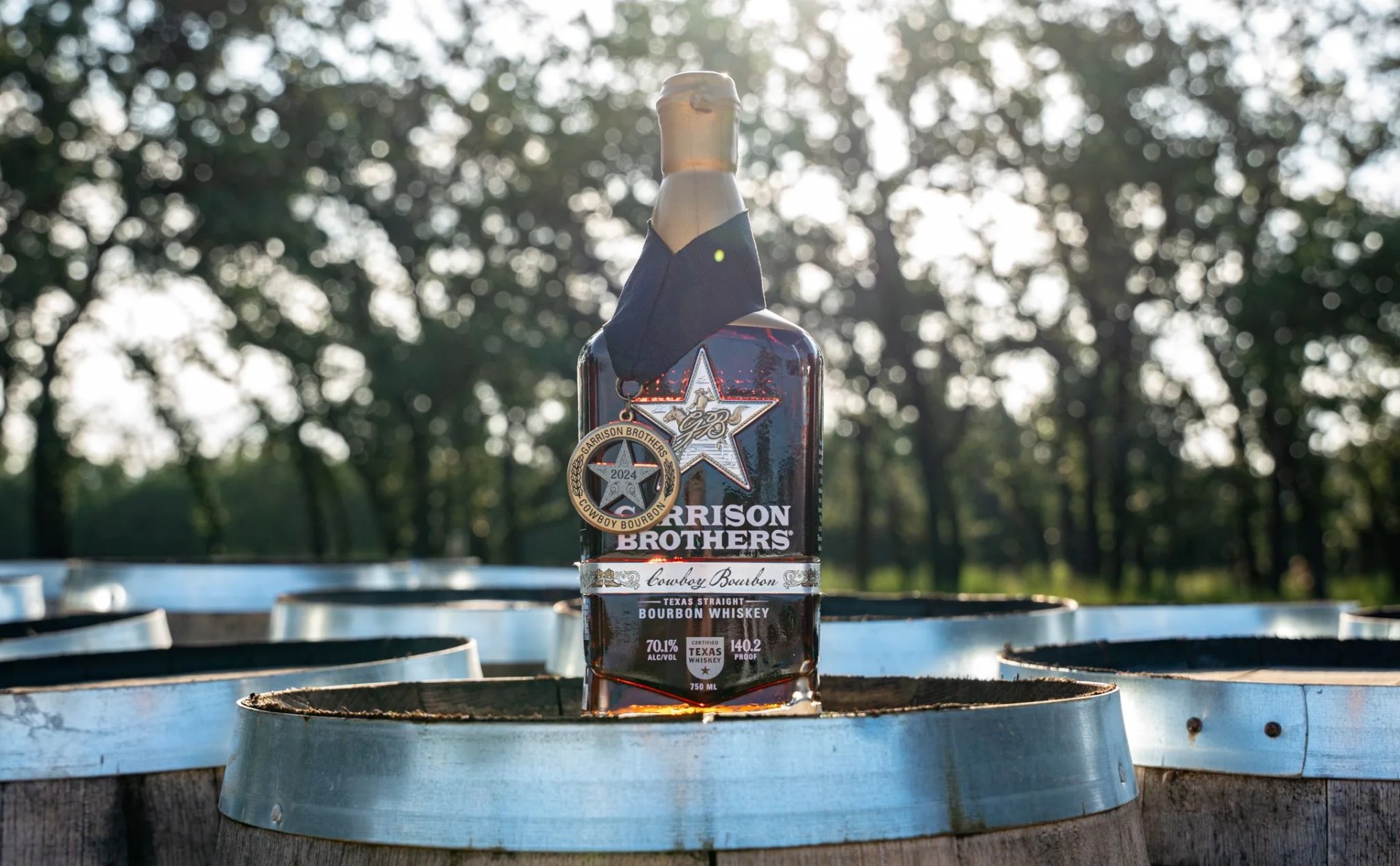 A bottle of Garrison Brothers Cowboy Bourbon 10 Year anniversary edition sitting on a bourbon barrel next to other bourbon barrels outside with a forest in the background.