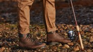 A person in khaki pants pictured below the knees wearing a pair of brown blundstone lace up boots standing on a rocky patch of ground holding a fishing pole