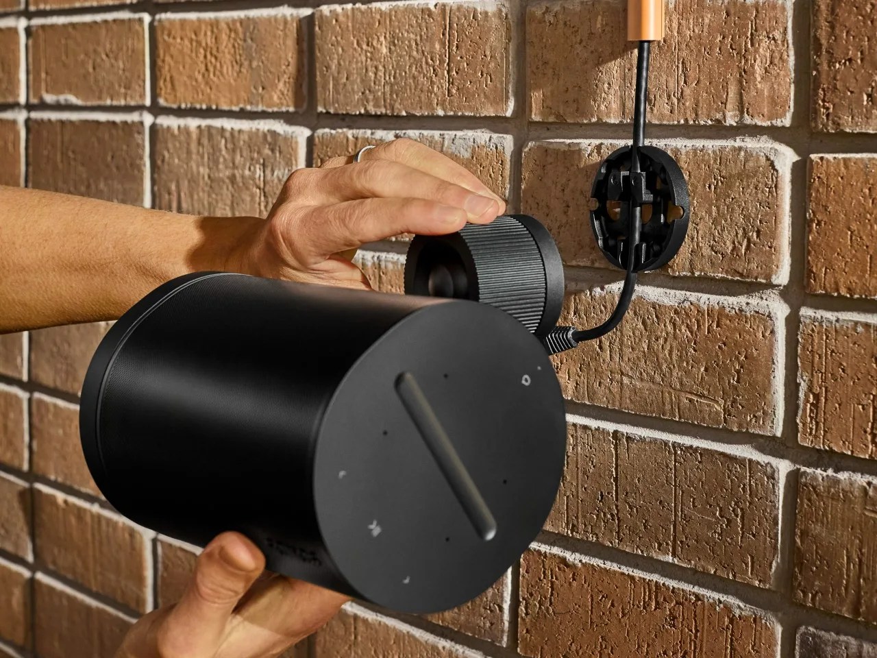 A black colored Sonos Era 100 Pro speaker being attached to its matching special wall mount attached to a brick wall.