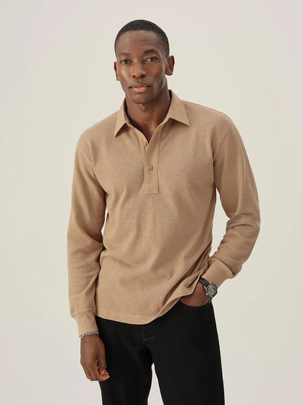 A black male model wearing Buck Mason&rsquo;s Field-Spec Rugby Polo in a Sandstone color that looks like a light tan. The model is facing forward with his hand in his pocket. 