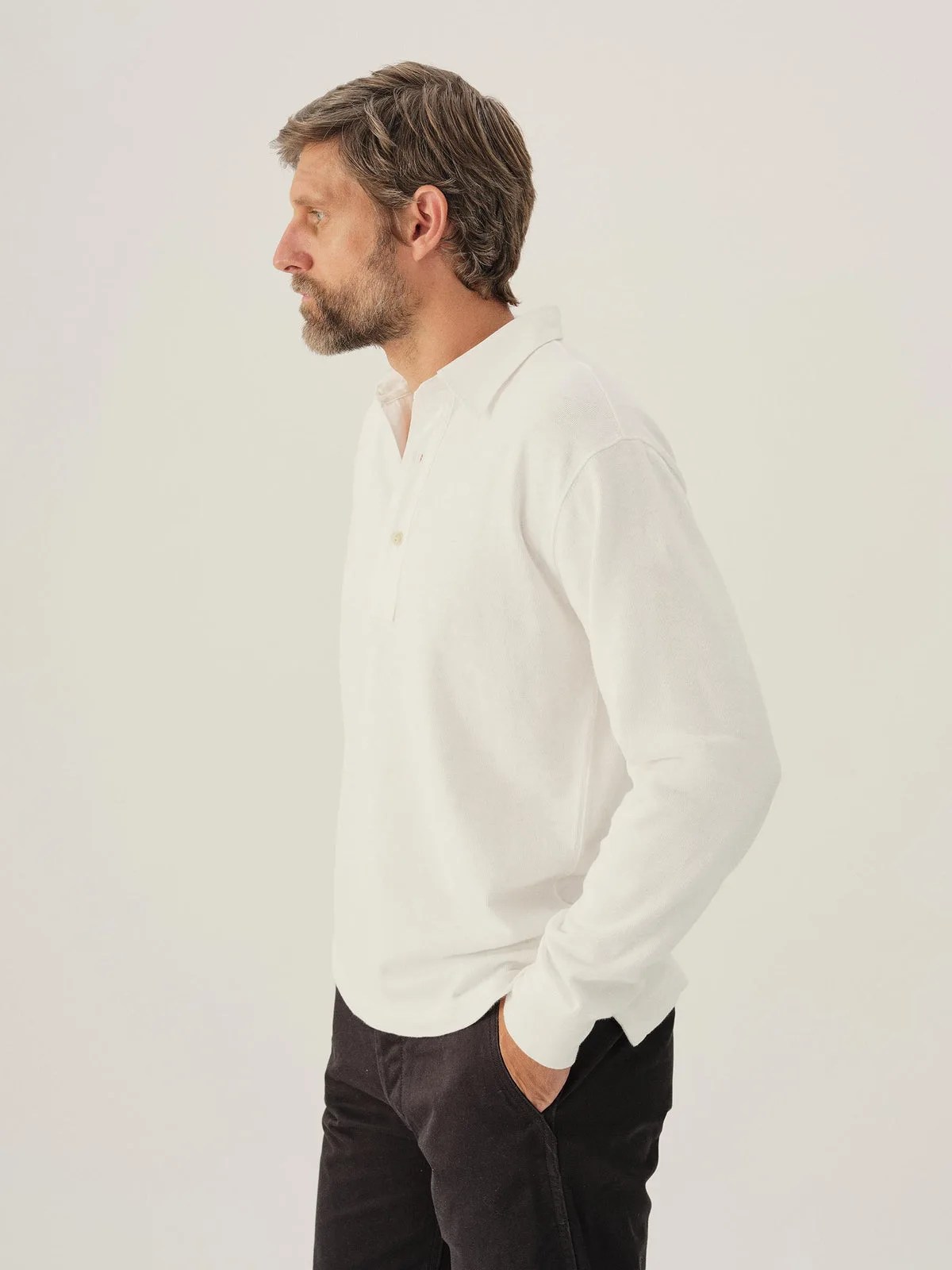 A white male model wearing Buck Mason&rsquo;s Field-Spec Rugby Polo in a white color. The model is facing to the left with his hand in his pocket. 