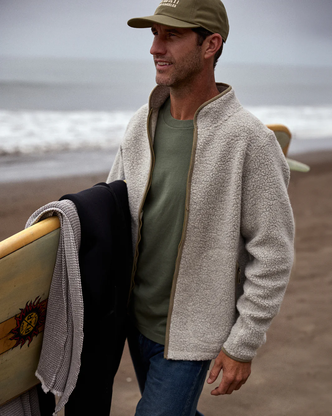 A man wearing an Outerknown Woolaroo Fleece jacket walking on a beach carrying a surfboard under one arm looking in the distance.