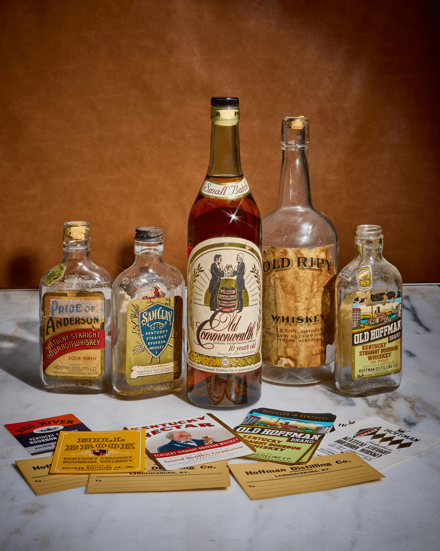 A vintage bottle of Old Commonwealth 10 Year Old bourbon flanked by empty vintage bottles of whiskey also distilled at the same distillery. The bottles are sitting on a marble countertop and backgrounded by a tan wall. Several cards showing bourbon labels are also laid out in front of the bourbon bottles.