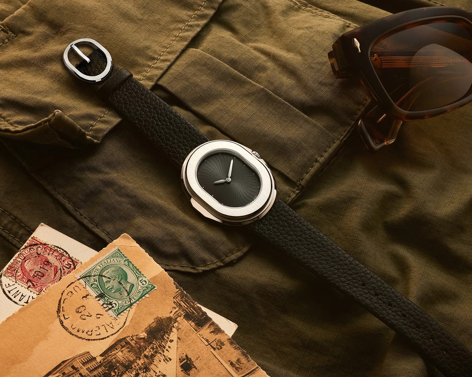 a serica dress watch on an olive safari shirt next to a pair of sunglasses and some postcards