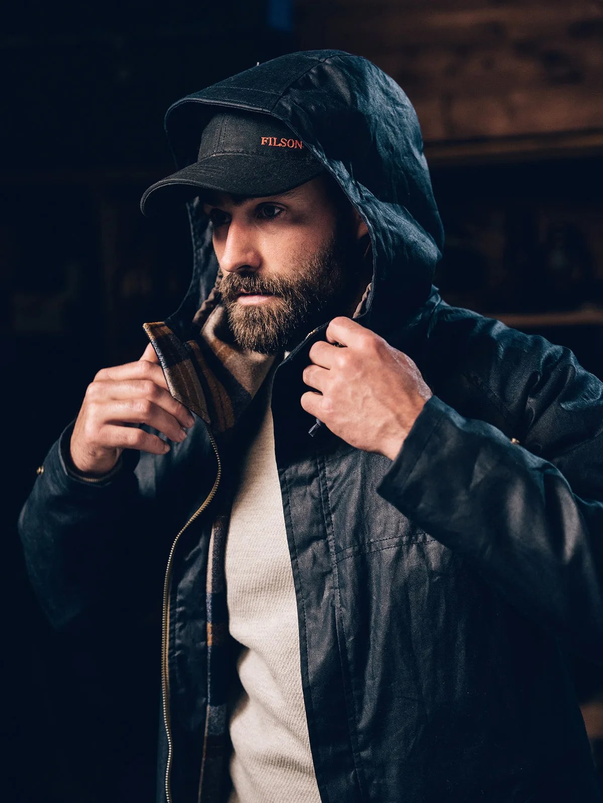 A man wearing the Filson x Freeman Rainjacket 2024 edition The man is wearing the jacket with the hood flipped up over a black baseball hat. His hands are shown pulling at the jacket hood's straps. The sweater beneath the jacket is oatmeal. Dark wood / brick walls appear blurred in the background. 