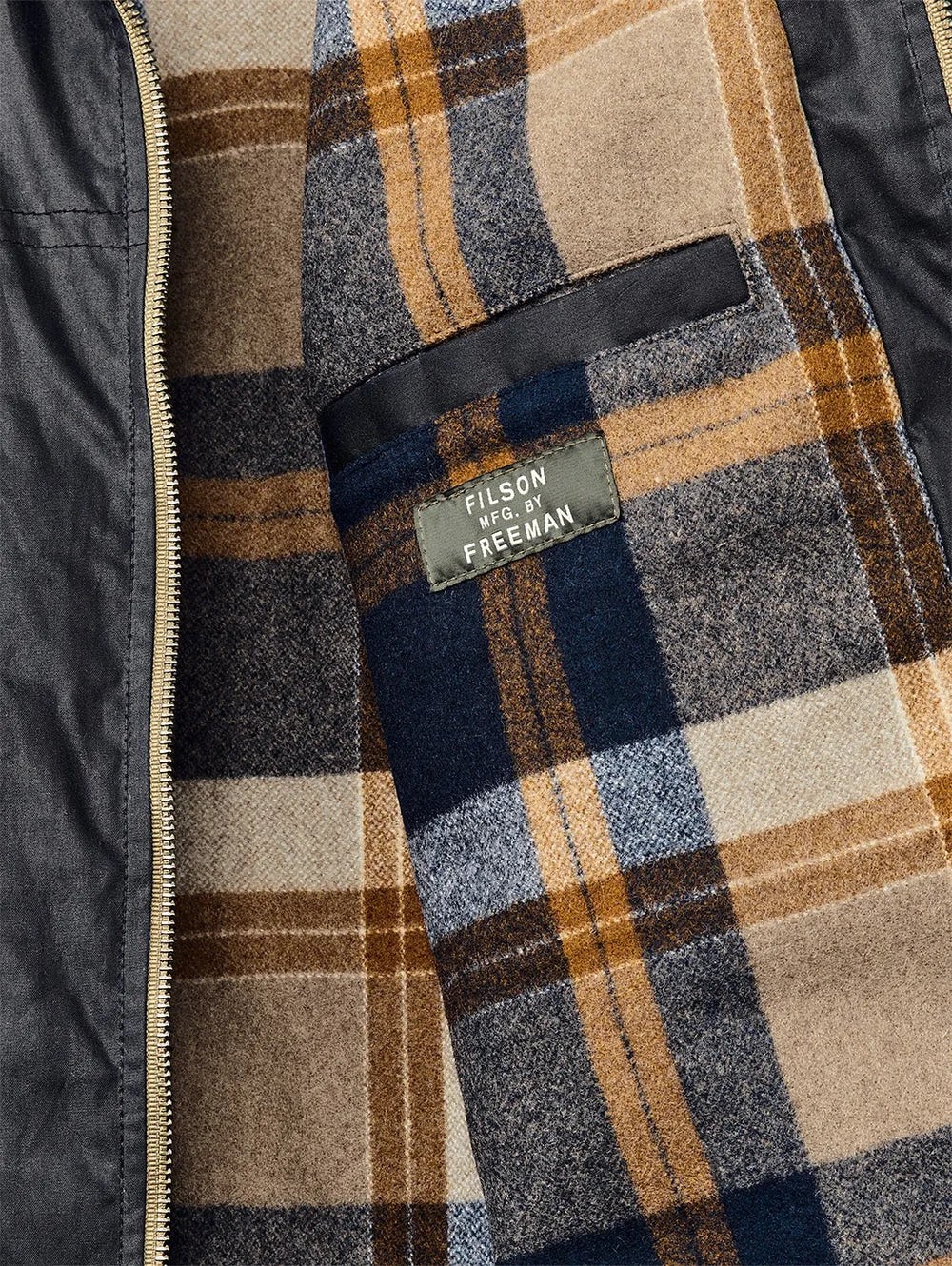 A close up look at the tag and flannel lining of the Filson x Freeman Wool Lined raincoat released in 2025. The image shows a closeup of the inner right flap of the jacket, highlighting the navy, gold and light tanned checked flannel pattern. 